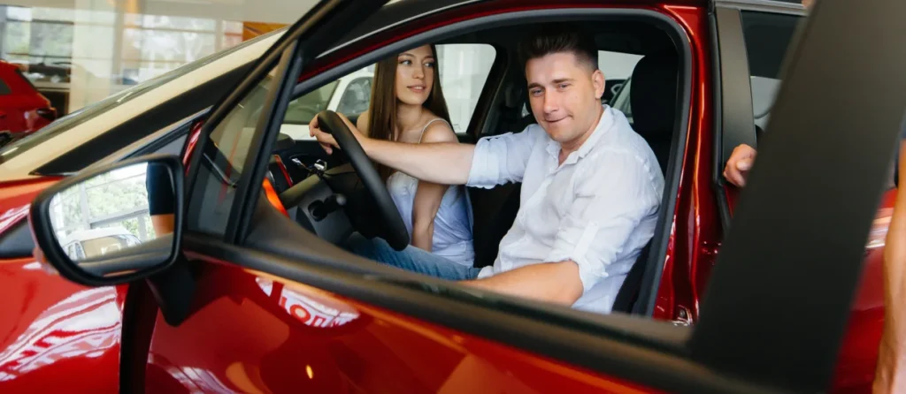 A smiling man and woman sitting in the front seats of a new red car in a showroom.