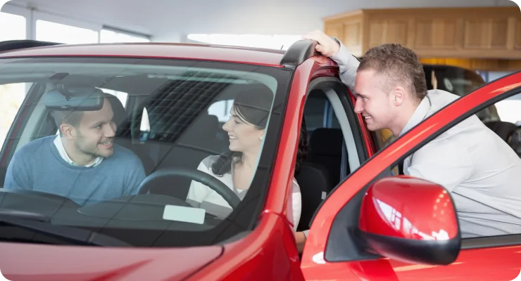A car salesman speaking with a male customer who is sitting in the driver's seat of a red car, likely discussing a warranty or purchase.