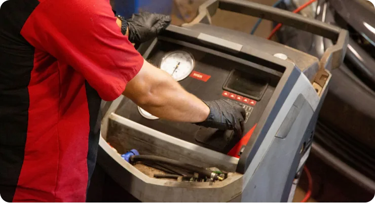 A close-up of a technician in a red shirt working on the air conditioning components in the engine bay of a car.