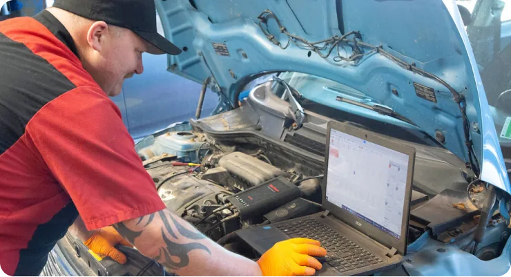 A mechanic in a red shirt looking under the open hood of a car while using a laptop or tablet for diagnostics.