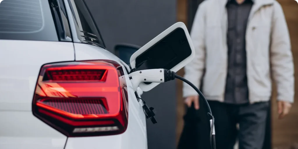 The rear view of a white electric or hybrid car being charged, with a charging cable plugged into its port and a person standing nearby.
