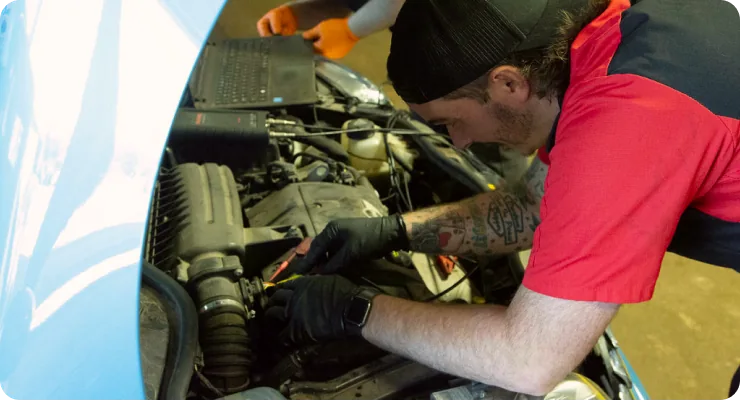 A technician in a red shirt leaning into the engine bay of a white car, performing maintenance or a visual inspection.