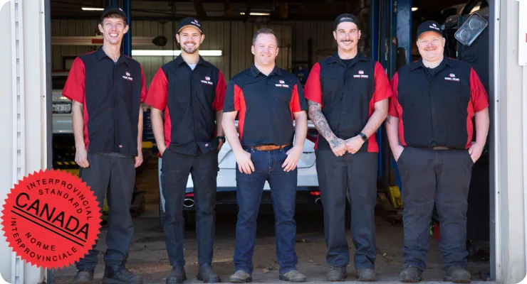 A team of five male mechanics wearing black and red uniforms standing proudly in a service bay, with a large Red Seal Canada Guaranteed Service logo overlaid on the image.