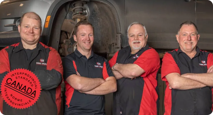 A group of four male mechanics in red and black work shirts, smiling and standing together in a garage, with a Red Seal logo visible on one shirt.