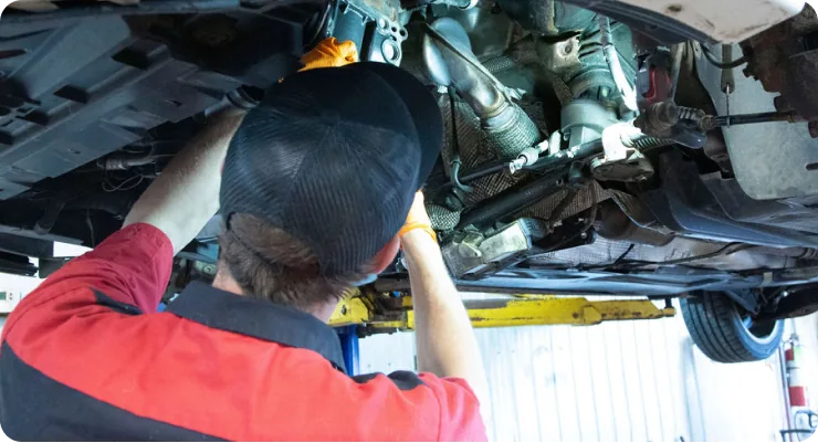 A mechanic in a red shirt under a raised car, working on the transmission or undercarriage components.