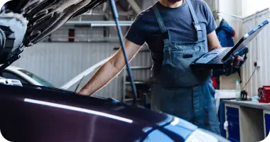 A mechanic in a blue uniform using a flashlight to inspect the inner workings of a car's engine.