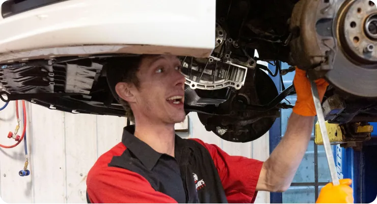 A smiling mechanic in a red and black uniform under a raised vehicle, inspecting the suspension or undercarriage.