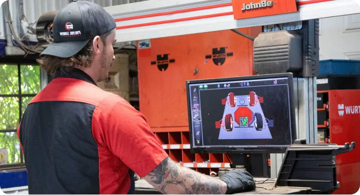A mechanic seated in a garage, looking at a computer monitor displaying a wheel alignment diagram and data.
