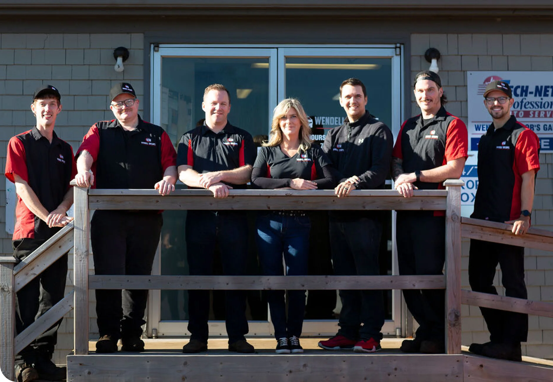 A group photo of six men and one woman, all staff members, wearing matching black and red work shirts, standing behind a wooden railing in front of the entrance to an auto service business. The building has light gray siding and large glass doors.