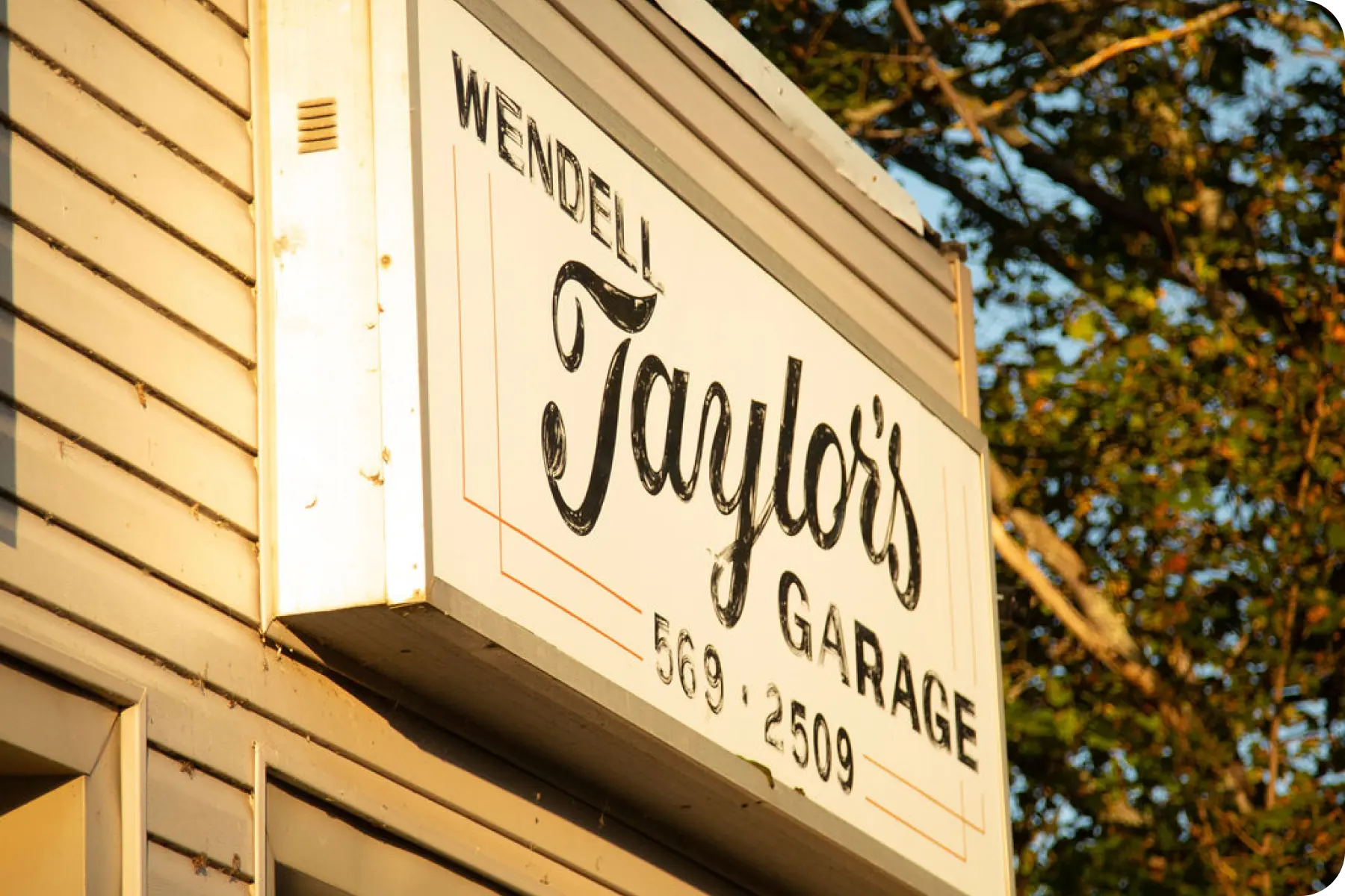A close-up of a lighted sign reading 'Wendell Taylor's Garage' against a light-colored wall and dark green trees.