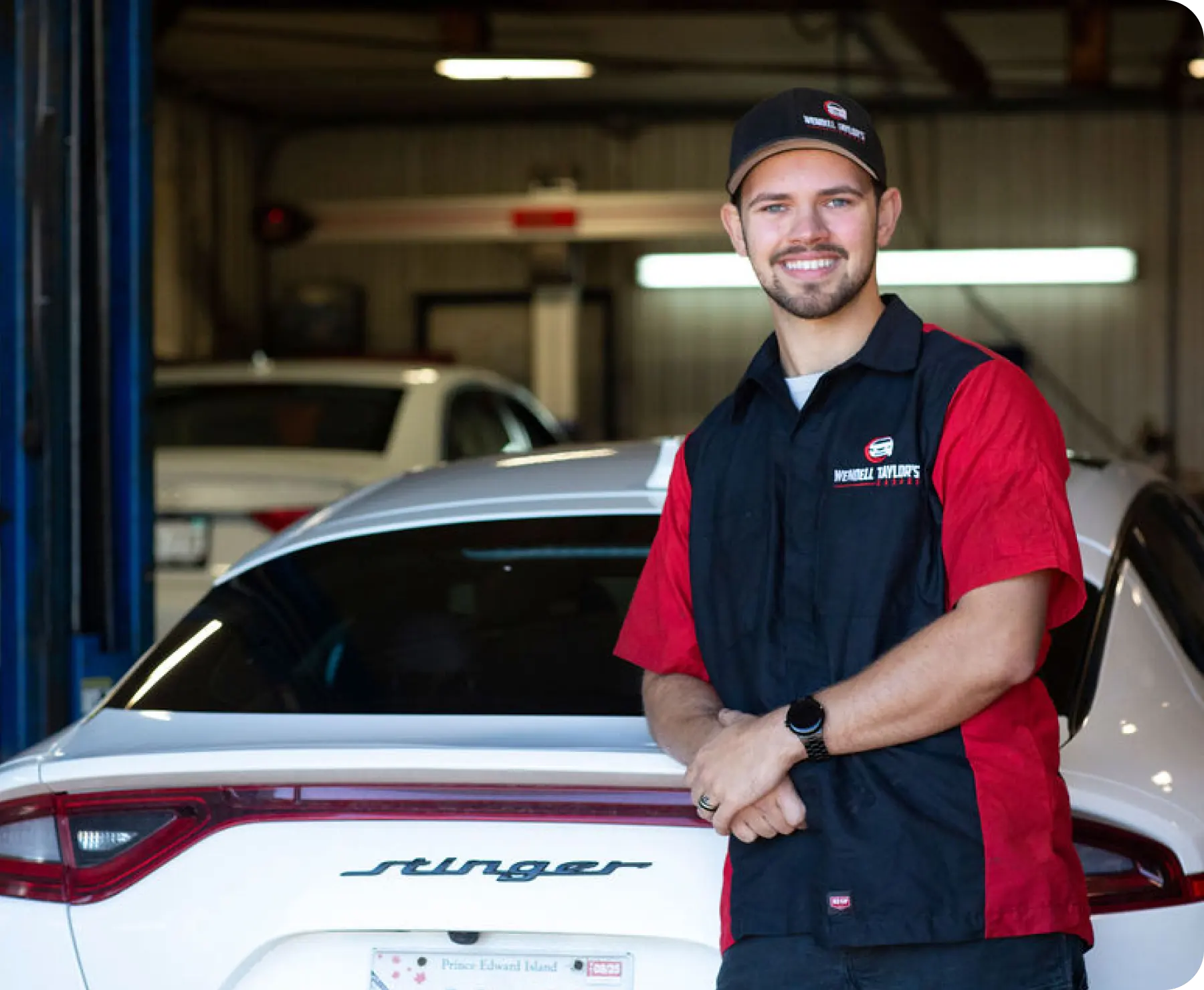 A smiling male mechanic in a red and black uniform standing in a service bay next to a white Kia Stinger sedan.