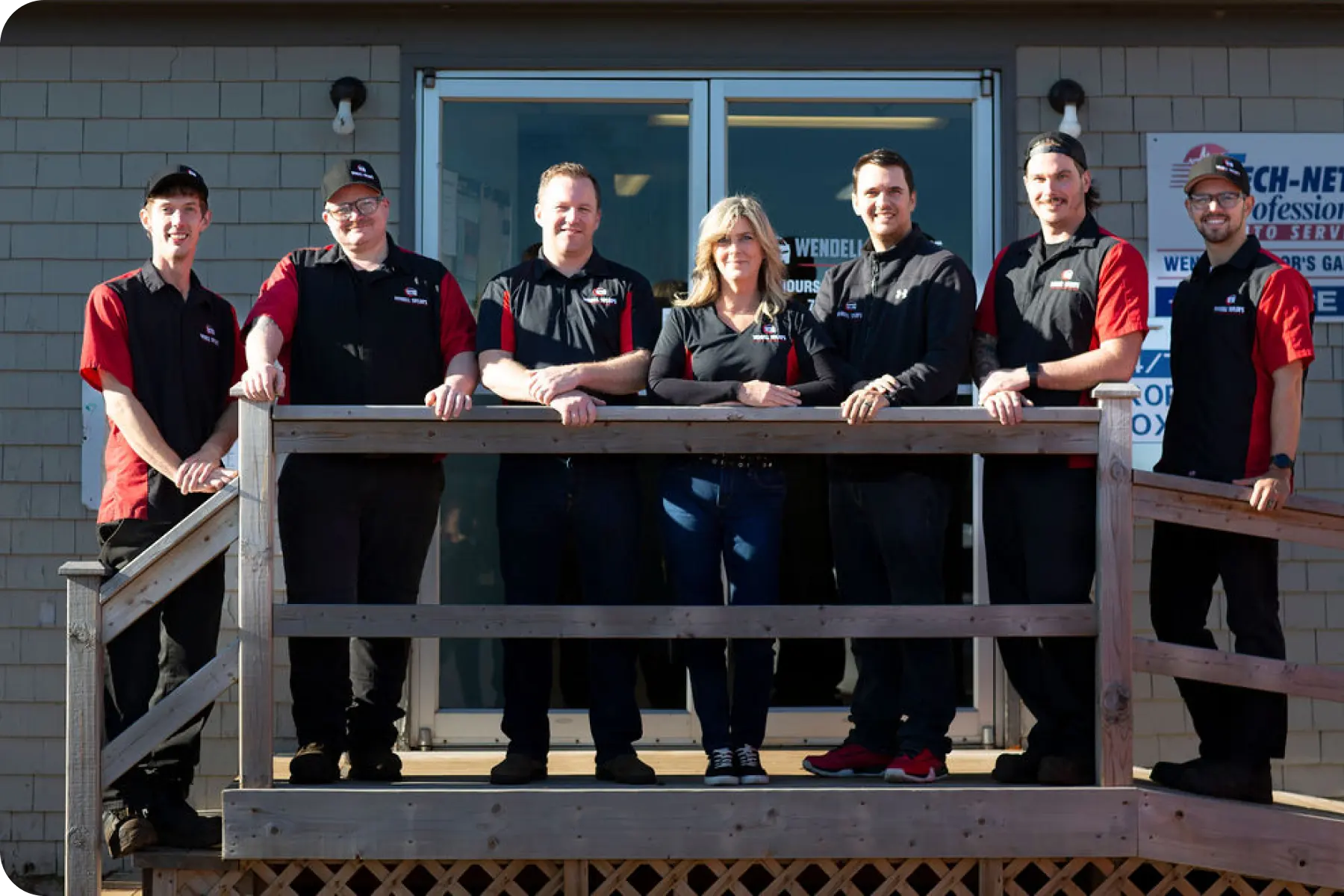 A group photo of six men and one woman, all staff members, wearing matching black and red work shirts, standing behind a wooden railing in front of the entrance to an auto service business. The building has light gray siding and large glass doors.