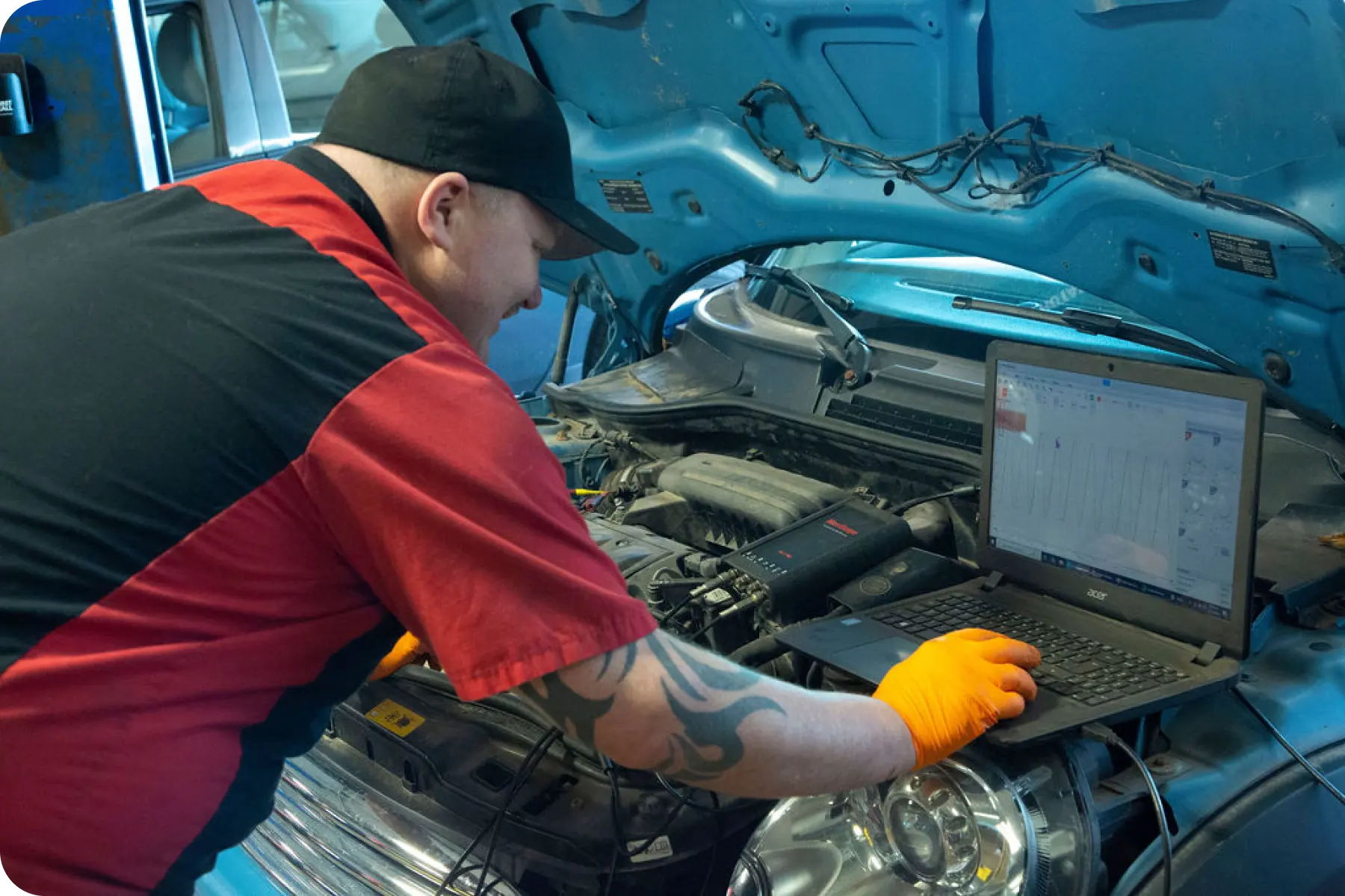 A mechanic in a red shirt looking under the open hood of a car while using a laptop or tablet for diagnostics.