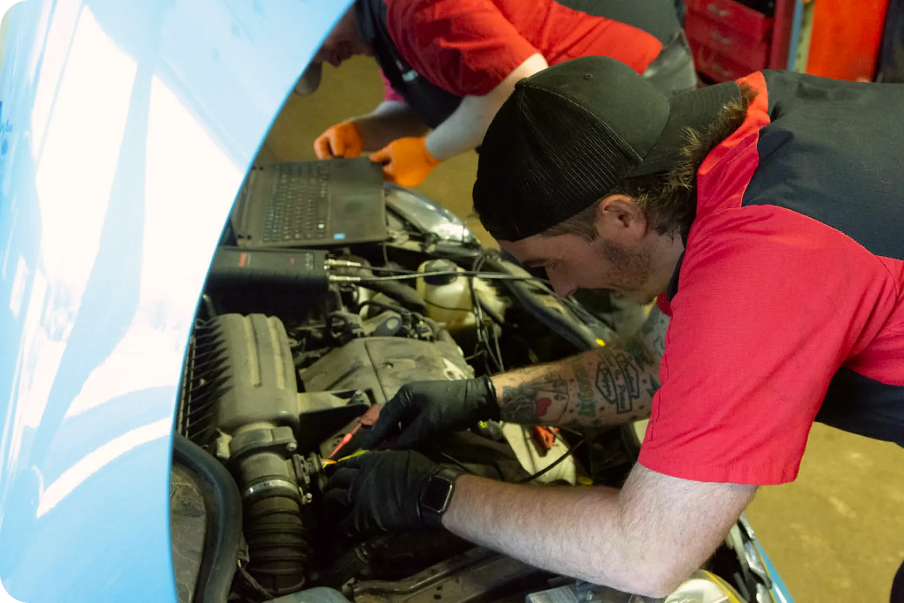 A technician in a red shirt leaning into the engine bay of a white car, performing maintenance or a visual inspection.