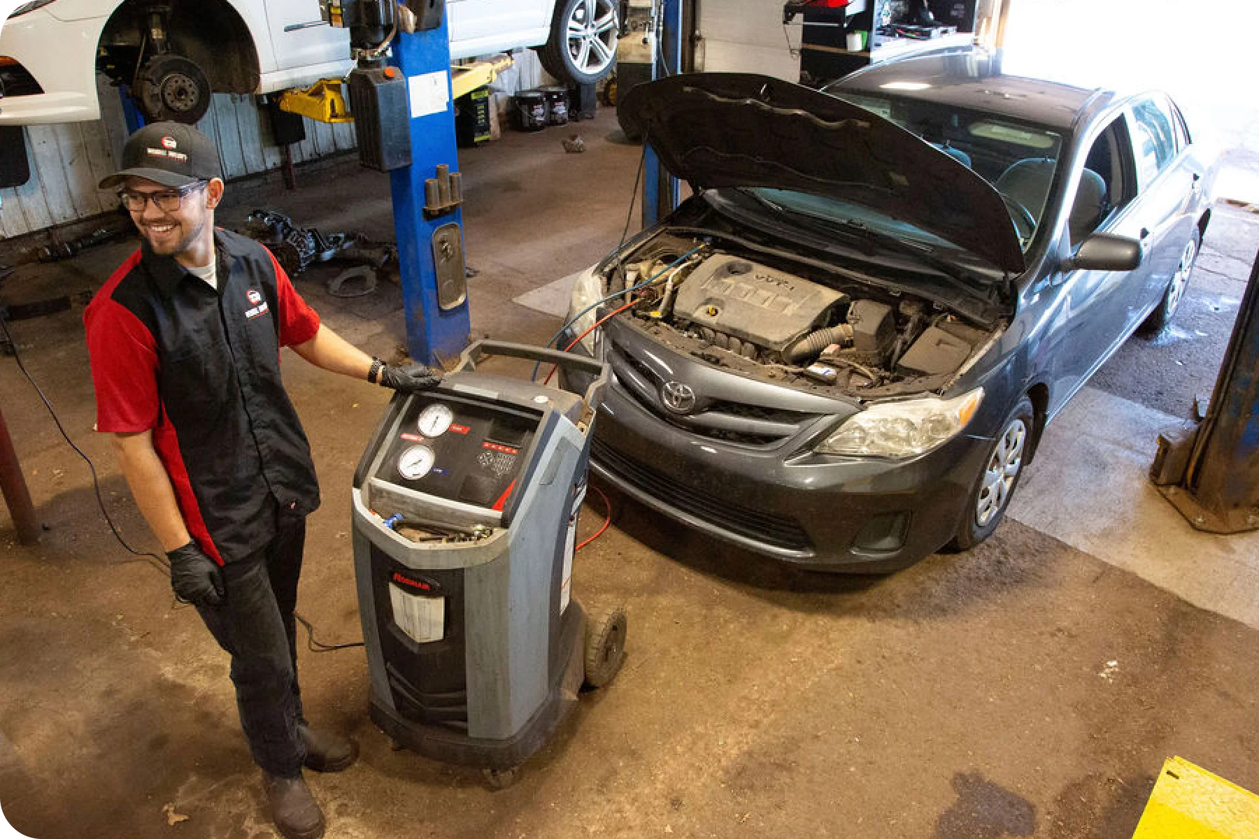 A smiling mechanic in a red and black uniform is standing next to an A/C service machine in a garage. A dark gray sedan with its hood open is visible next to the machine, ready for service.