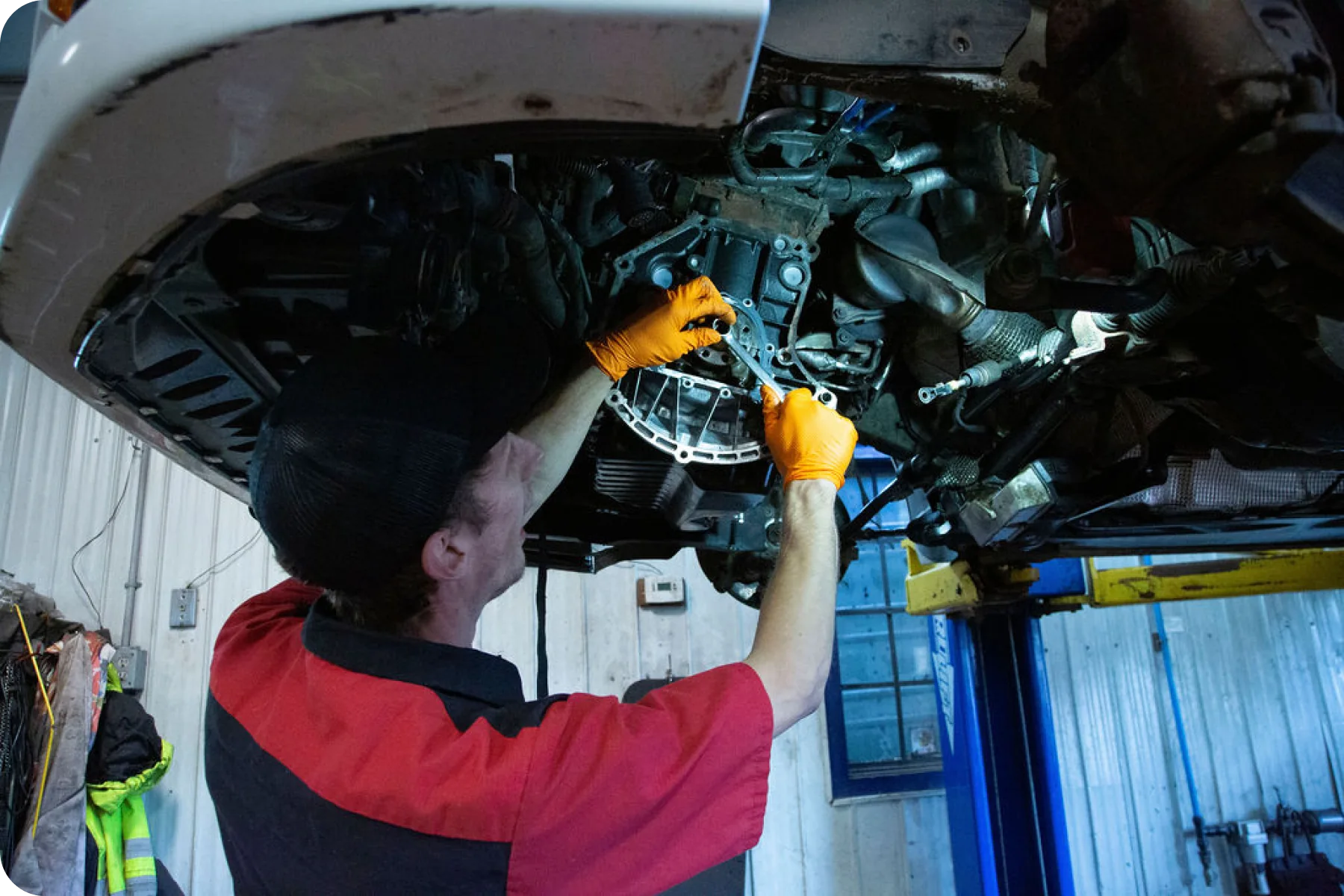 A mechanic in a red and black shirt and a baseball cap works on the underside of a car, illuminated by a bright light.