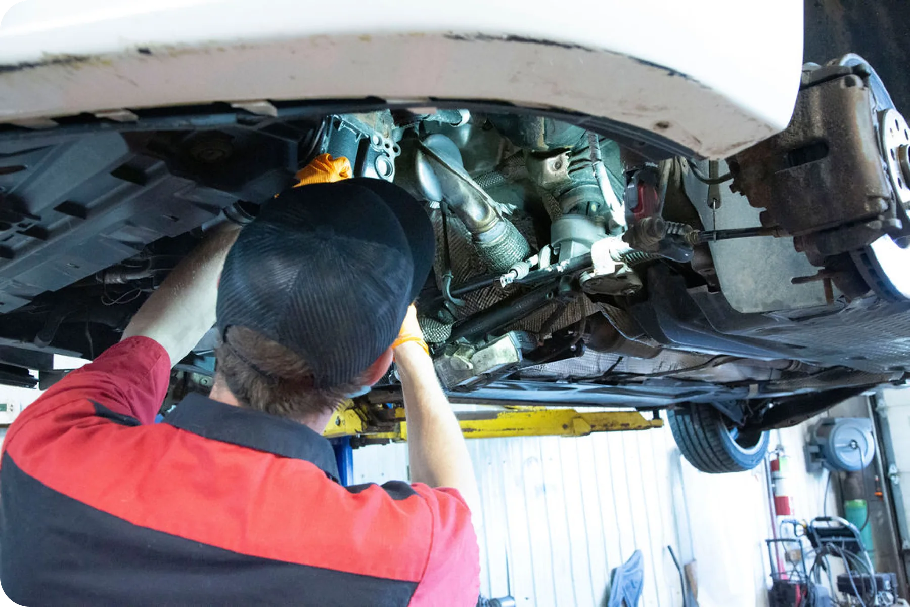 A mechanic in a red shirt under a raised car, working on the transmission or undercarriage components.