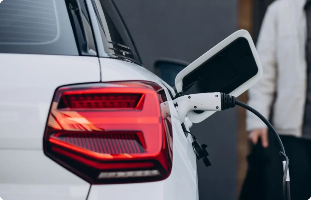 The rear view of a white electric vehicle being charged, showing the illuminated taillight and the charging cable plugged into the port.
