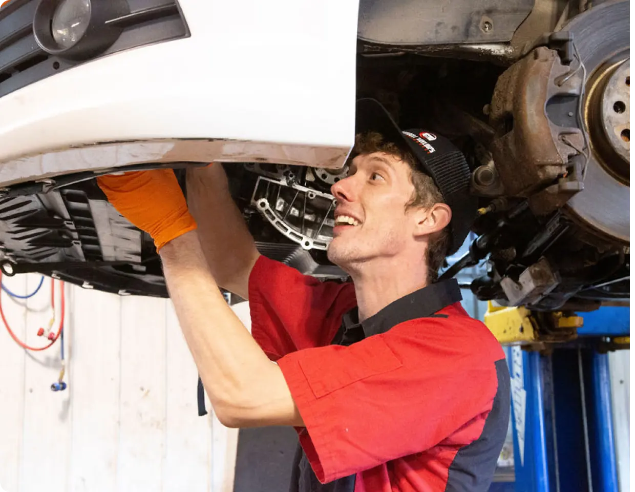 A mechanic in a red shirt and baseball cap, wearing orange safety gloves, smiling while working on the undercarriage or front bumper of a white vehicle on a lift.