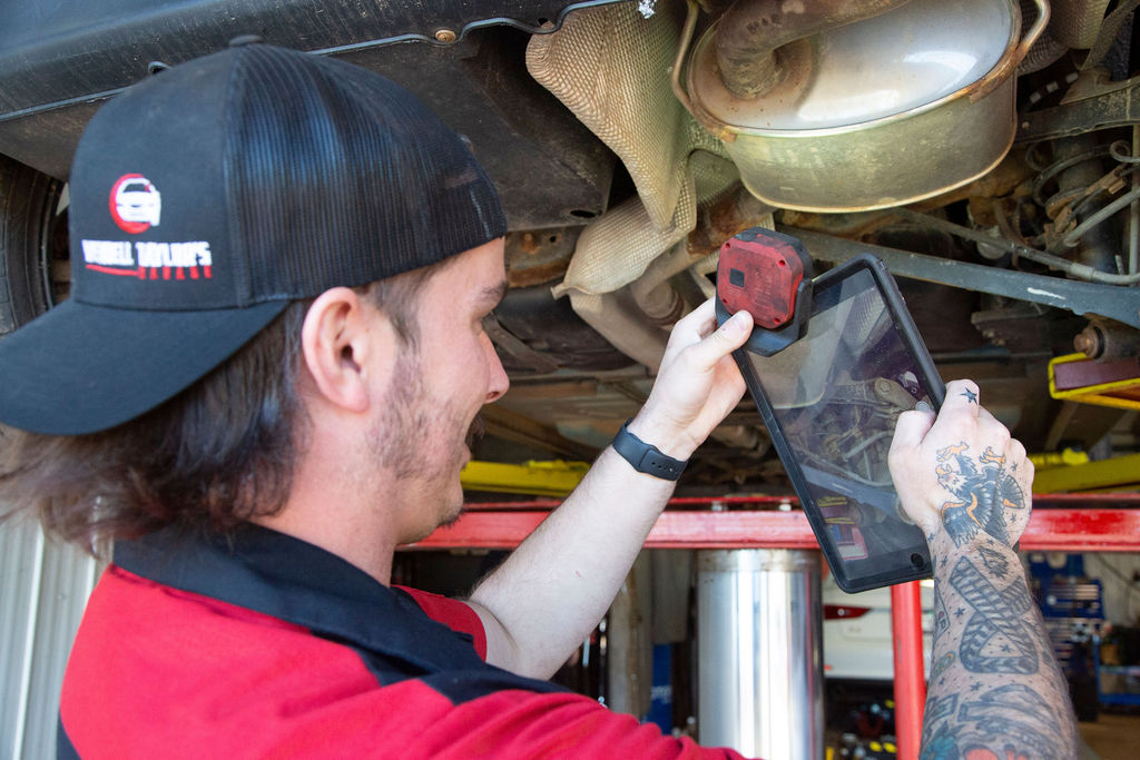 A mechanic in a red shirt and black hat working underneath a vehicle raised on a lift, wearing gloves.