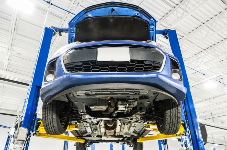 A blue car is elevated high on a yellow hydraulic lift inside a bright, clean auto repair garage, viewed from the ground looking up at the exposed undercarriage.